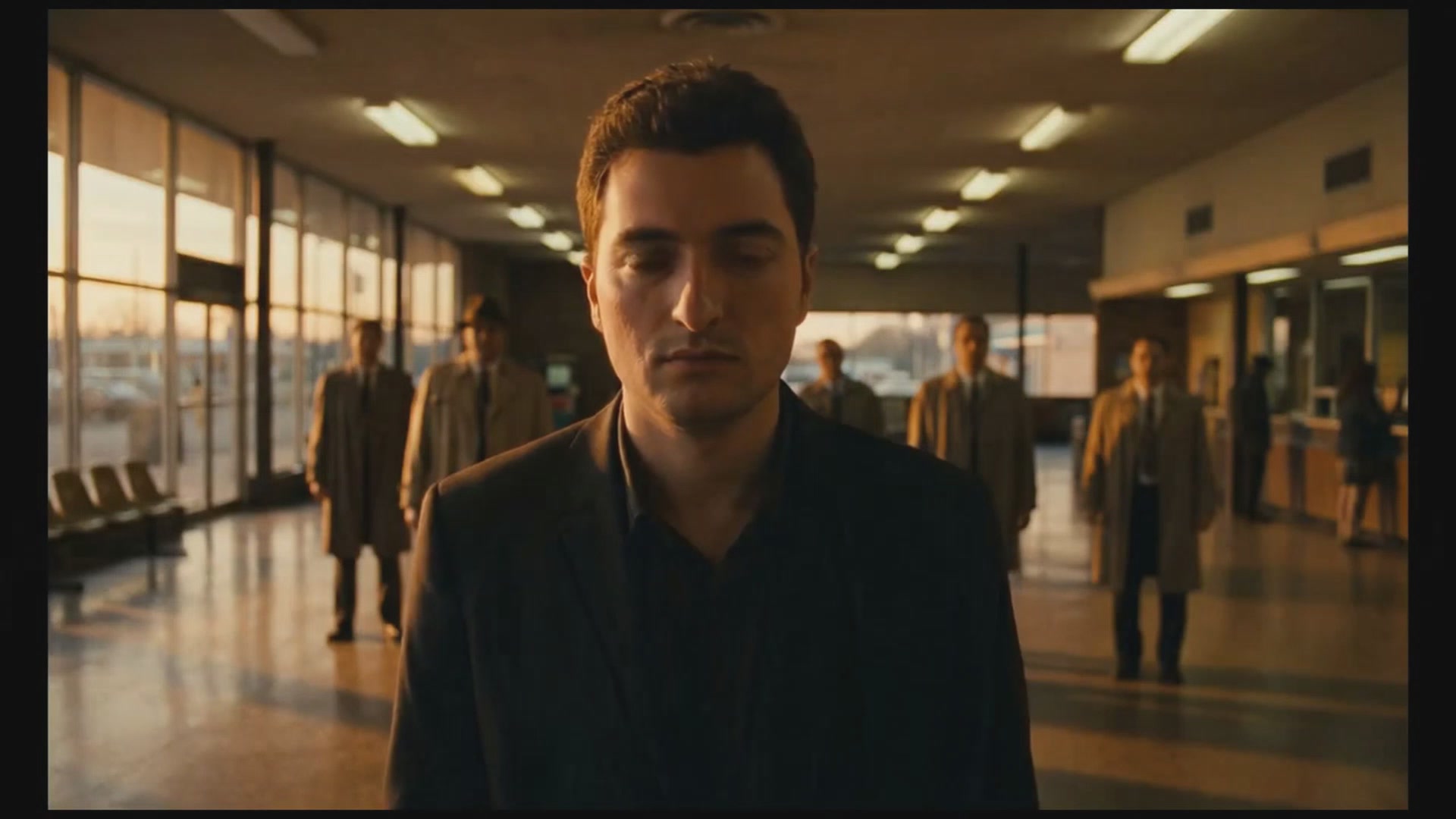 The image shows a man wearing a suit standing alone in an airport terminal. The scene includes several other people walking around or sitting at various points within the terminal. There are also chairs visible, suggesting that this could be a waiting area for passengers.