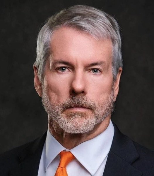 A portrait of a gray-haired, bearded man wearing a dark suit, light blue shirt, and orange tie against a dark background.
