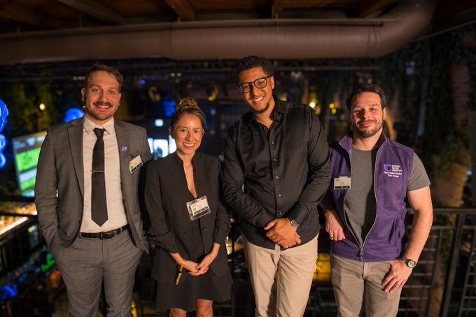 Four people posing for a photo at what appears to be a tech or gaming event, with one man on the right wearing a purple vest labeled 'One Step Closer'.