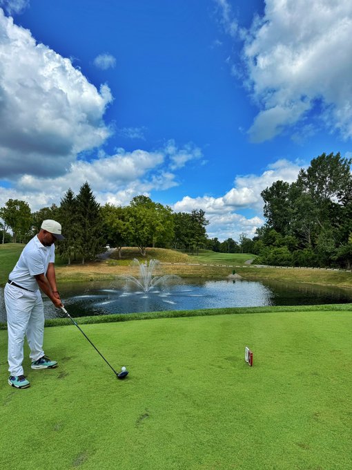 A golfer wearing a white cap, white shirt, and white pants tees off on a lush green course with a picturesque pond and fountain in the background, pairing perfectly with the relaxed "Time to golf" sentiment after waiting out a crypto pullback.
