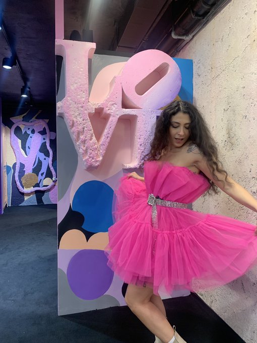A woman with long curly hair wearing a bright pink strapless tulle dress posing in front of a large 3D 'LOVE' sculpture in pink and blue tones.