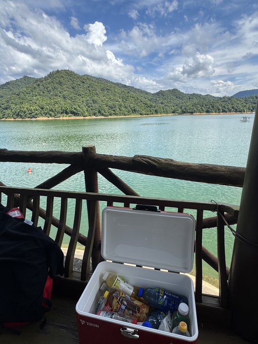 An open cooler labeled 'TRION' filled with drinks and snacks sits on a wooden deck overlooking a scenic lake with green mountains in the background.