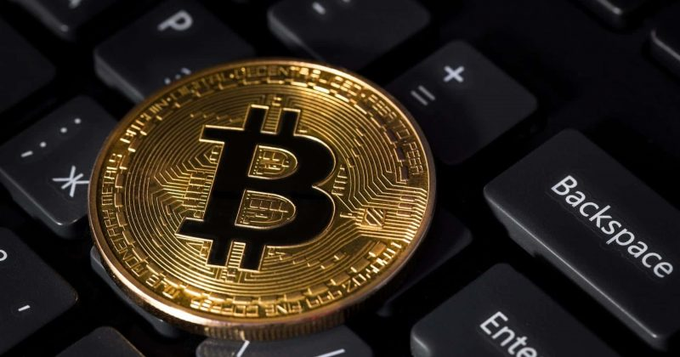 A close-up photograph displays a gold-colored Bitcoin physical coin resting on a black computer keyboard, with the Backspace and Enter keys visible.