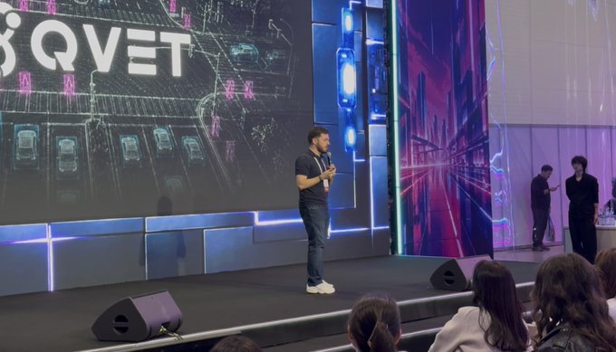 A man giving a presentation on stage in front of a screen displaying the 'QVET' logo, with attendees visible in the audience.