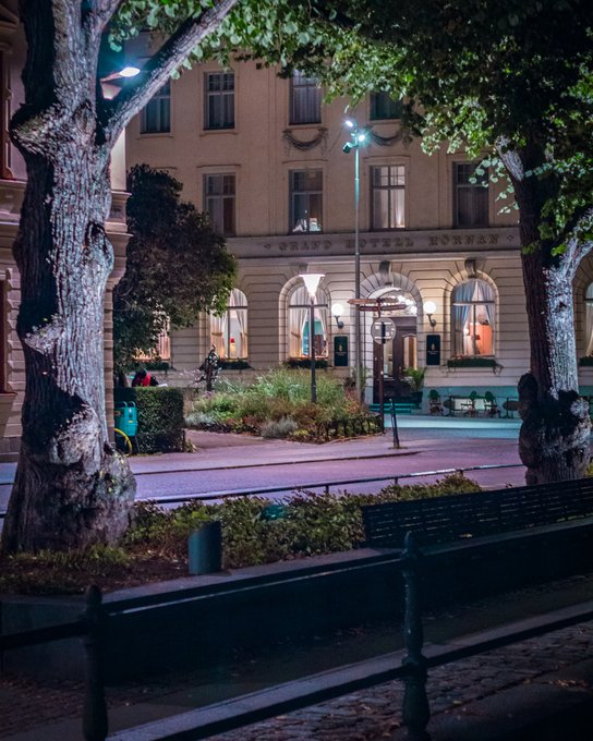 A nighttime view of the 'Grand Hotel Hornan' building, seen past a tree-lined street with a bench in the foreground.