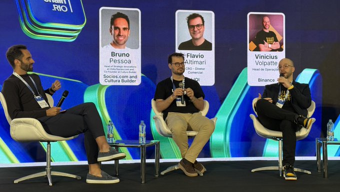 Three men sit on white chairs on a stage at Blockchain Rio, with a screen behind them displaying headshots and titles for Bruno Pessoa (Head of Strategy, Sócios.com and Cultura Builder), Flavio Altimari (CEO, Itaú Commercial), and Vinicius Volpatte (Head of Operations, Binance), as Volpatte speaks into a microphone during his panel on "AI + RWA: artificial intelligence and real-world assets on-chain."