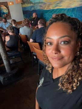 A selfie of a woman with long curly highlighted hair wearing a dark t-shirt, taken in what appears to be a restaurant or bar with other patrons seated at tables in the background.