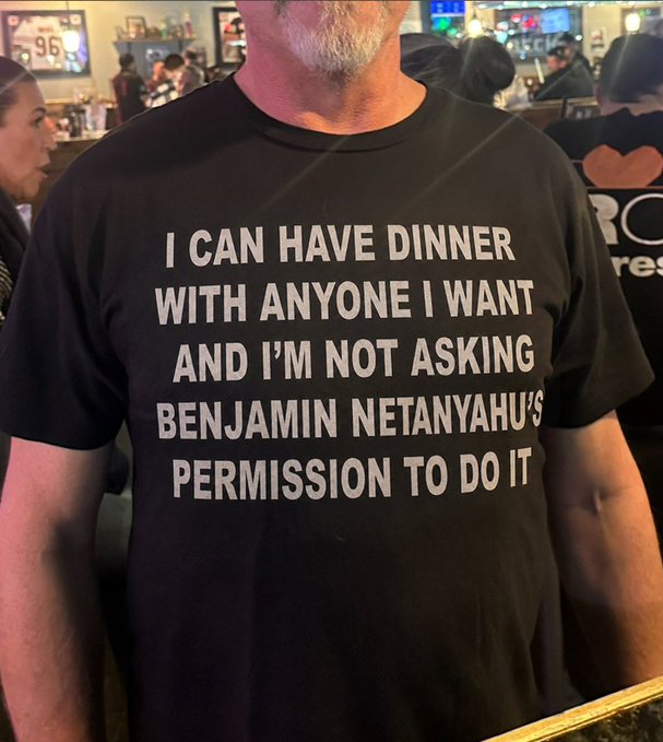 A man wears a black t-shirt with the text 'I CAN HAVE DINNER WITH ANYONE I WANT AND I'M NOT ASKING BENJAMIN NETANYAHU'S PERMISSION TO DO IT'.