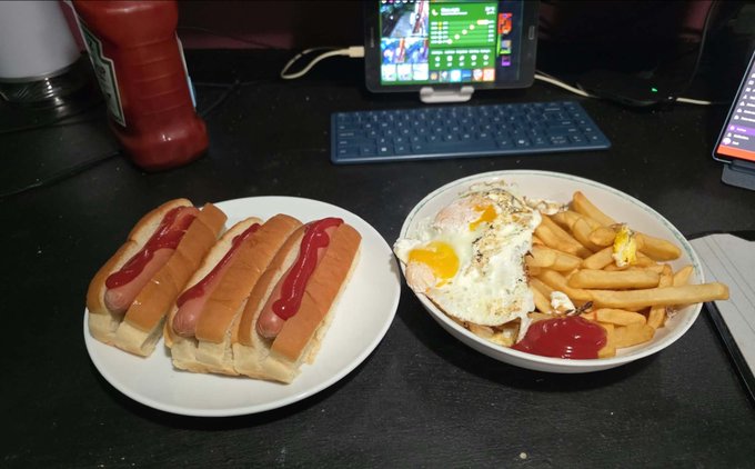 A meal consisting of three hot dogs with ketchup on a white plate alongside a bowl of fries and a fried egg, set on a black table near a tablet displaying what appears to be a football broadcast.