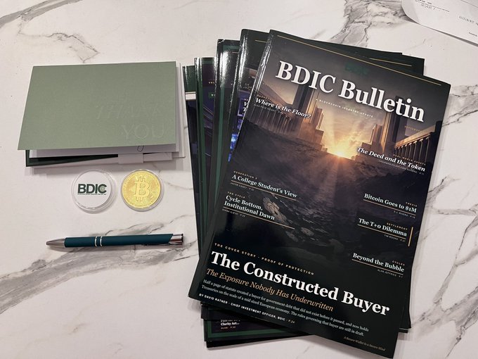A stack of 'BDIC Bulletin' newspapers with cryptocurrency-themed headlines like 'The Constructed Buyers' and 'Bitcoin Goes to $8M', displayed alongside a 'THANK YOU' card, pen, and Bitcoin-themed coins on a marble surface.