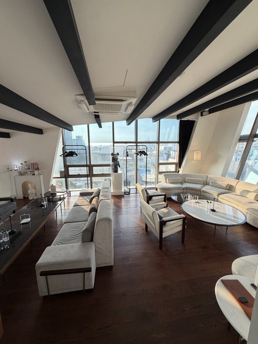 A spacious modern living room with exposed dark ceiling beams, featuring a long white sectional sofa, armchairs, marble coffee tables, sculptural artwork, and floor-to-ceiling windows overlooking a cityscape.