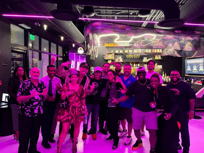 A group of people posing for a photo inside a bar or club with neon pink lighting, including one person dressed in drag, with a 'Collectors Inn' sign visible in the background.