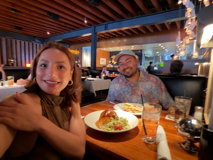 A couple smiling at a restaurant table, with plates of food including fries and guacamole, glasses of water, and a dimly lit, modern interior in the background.