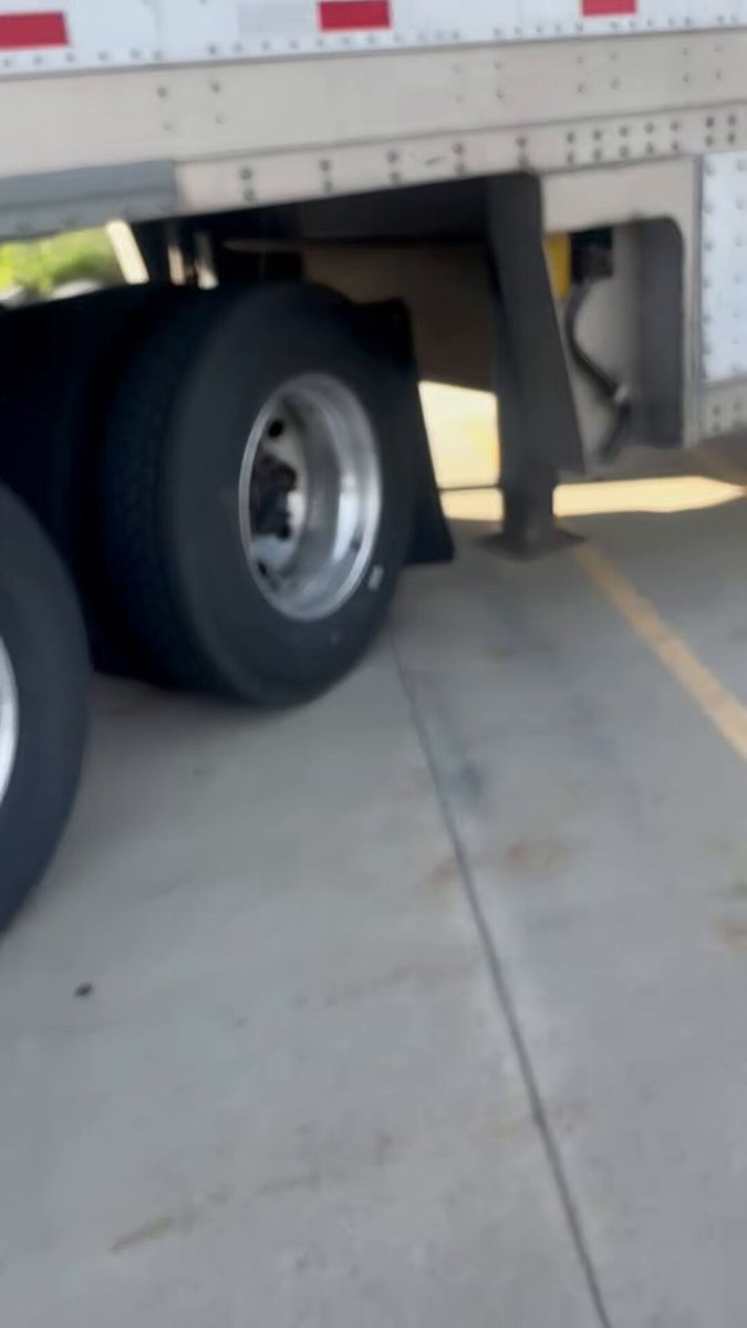 The image shows a pair of black tires with red reflectors installed under a semi-truck, parked on a concrete surface indoors like a warehouse or a storage area for trucks. A yellow tag can be seen hanging from one of the tires. Below the tires are some brown boxes and a white floor. There is also a text that reads " Almost Friday" followed by the word "DOG", which might be related to dog food sales or promotions on Fridays, coinciding with the truck driver's weekend off. The image highlights the tire replacement process for trucks during the week when dog food sales are typically less active due to the holiday weekend.
