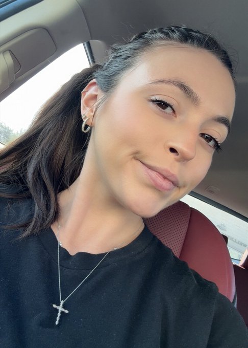 A selfie of a smiling young woman with long dark hair, wearing a black shirt and a silver cross necklace, seated inside a car.
