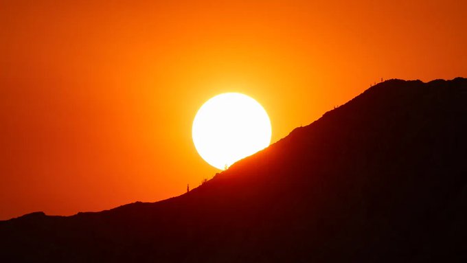 A bright orange sun setting behind the dark silhouette of a mountain ridge, with two tiny silhouettes of people visible on the peak.