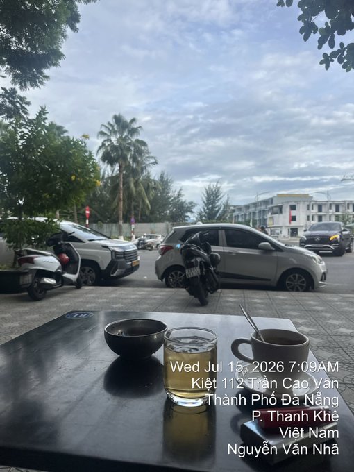 A view from inside a café in Đà Nẵng, Vietnam showing a table with a bowl, glass of water, and coffee cup, with a street scene featuring parked cars, motorcycles, palm trees, and a traffic light. Text reads: 'Wed Jul 15, 2026 7:09AM, Kiệt 112 Trần Cao Vân, Thành Phố Đa Nẵng, P. Thanh Khê, Việt Nam, Nguyễn Văn Nhã.'