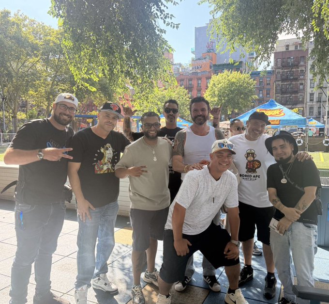 A group of about eight men posing together outdoors in a sunny urban plaza with trees and colorful umbrellas in the background.