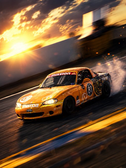 A yellow Mazda race car with the number 81 speeds along a track, kicking up smoke as it drifts against a bright sunset.