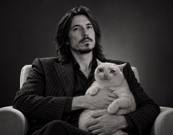 A black-and-white portrait of a man with long hair holding a white cat in his lap.