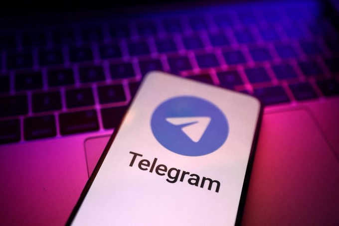 A smartphone displays the Telegram logo and app name, resting on a laptop keyboard illuminated by purple and pink lighting.