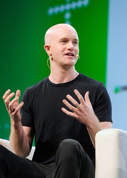 A bald man in a black t-shirt speaks while gesturing with both hands at a conference.