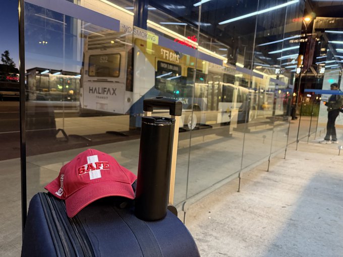 A blue suitcase sits at a bus station with a red 'SAFE' cap and a black tumbler on top, with a Halifax Transit bus visible behind the glass.