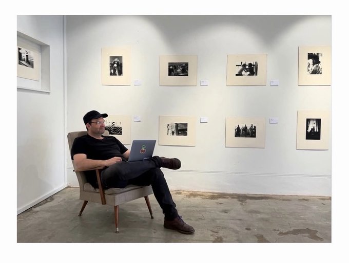 A man in a black cap and t-shirt sits in a chair with a laptop, photographed in an art gallery displaying black-and-white prints.