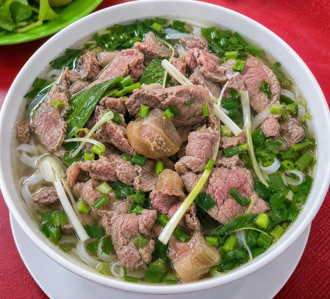 A white bowl filled with Vietnamese pho noodle soup, featuring sliced beef, green onions, cilantro, and broth, with a side plate of fresh herbs on a red tablecloth.
