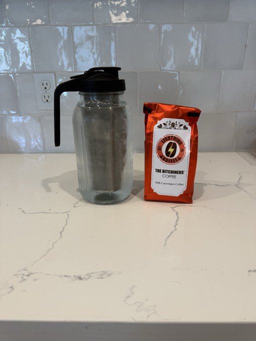 A glass beverage dispenser with a black lid sits on a white marble-patterned countertop next to an orange bag of 'The Ritchens Coffee' 100% Colombian Coffee.