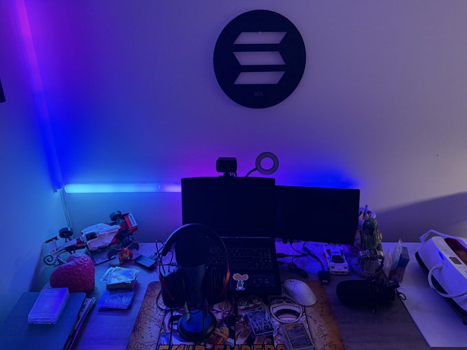 A dimly lit gaming/streaming setup illuminated by blue and purple LED strip lights, featuring a laptop, dual monitors, a webcam with ring light, headphones, and a Solana logo mounted on the wall.