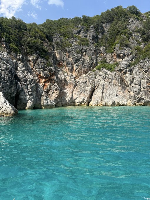 A scenic coastal view showing clear turquoise water meeting a rocky cliff face covered with green pine trees under a bright sky.