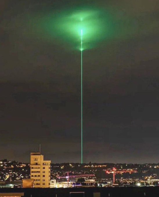 A nighttime cityscape featuring a prominent green laser beam shooting straight up into the sky above illuminated buildings and construction cranes.