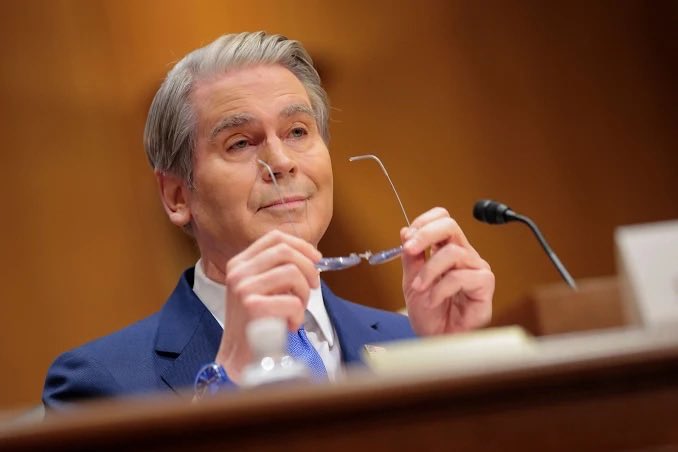 An older man in a blue suit sits at what appears to be a Senate hearing, holding up his eyeglasses while looking forward, with a microphone visible nearby.