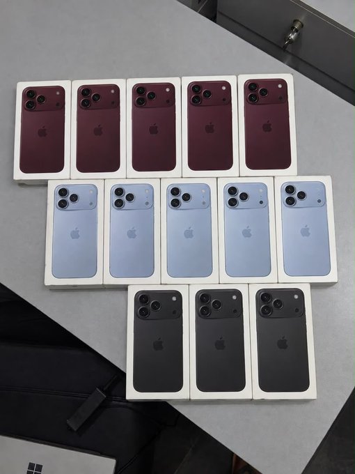 Thirteen sealed Apple iPhone Pro Max boxes are arranged in three rows—five in burgundy/red, five in light blue, and three in black—displayed on a gray surface as visual proof of the promised giveaway.