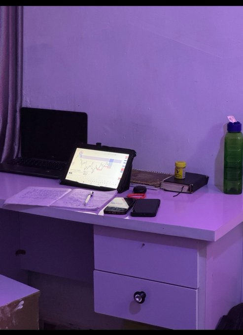 A trader's desk setup with a tablet displaying candlestick charts, a laptop, notebooks, and a green water bottle, all illuminated by purple lighting—perfectly capturing the "GM & happy new week, traders" trading-ready vibe.