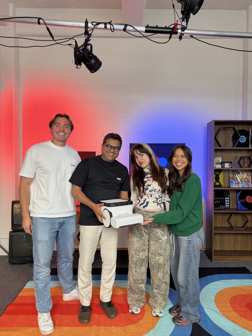 Four people stand in a studio with red and blue lighting, holding a PlayStation 5 console together; the setup includes a camera rig above and a colorful circular rug.