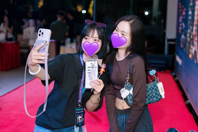 Two young women taking a selfie at an event, their faces obscured by purple heart emojis; one wears a 'VIRTUAL ON TOP' badge and the other carries a Coach bag.