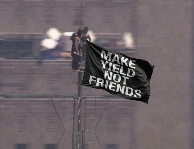 Two people stand on a rooftop holding a large black flag with bold white text reading 'MAKE YIELD NOT FRIENDS'.