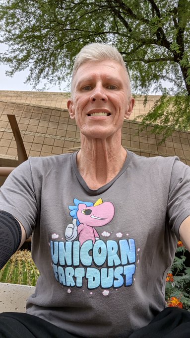 A man with short blonde hair wearing a gray t-shirt featuring a pink cartoon unicorn and the text 'UNICORN FART DUST', taking a photo outdoors with a building and tree visible in the background.