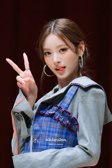 A young woman in a gray outfit and blue plaid overalls poses with a peace sign; the overalls read “shizuka” and “YOSHIKAWA.”