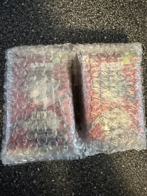 Two items wrapped in clear bubble wrap rest on a dark speckled surface, with partially visible red and yellow packaging underneath the plastic.