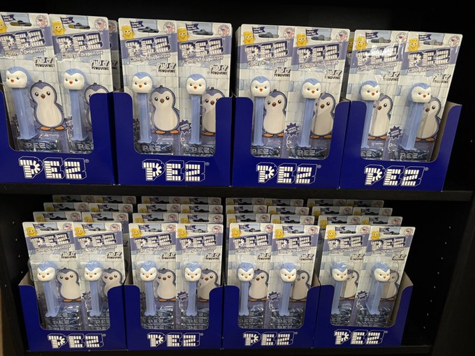 A retail store shelf displays neatly arranged merchandise featuring Pudgy Penguins characters on what appear to be PEZ candy dispensers, packaged in blue boxes labeled "PEZ" with Japanese text and "PENGU" branding. The image illustrates the retail product presence tied to the $PENGU token associated with the Pudgy Penguins NFT collection.