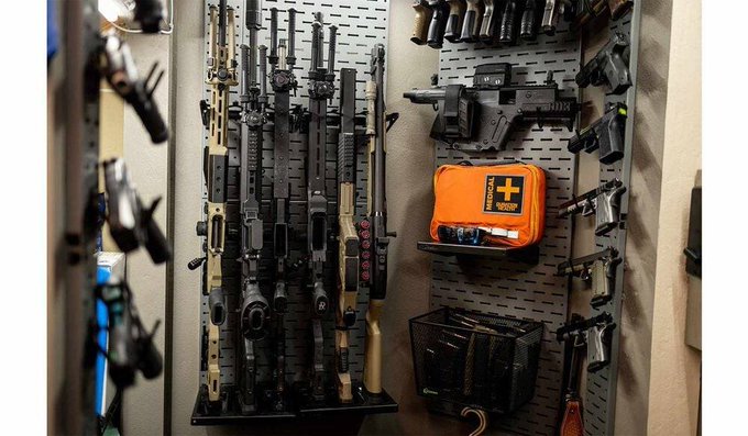 A wall-mounted gun safe or display case containing several rifles, handguns, and an orange 'RESCUE' medical pouch on a perforated metal backboard.