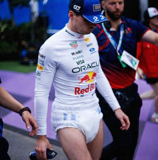 A Formula 1 driver wearing a Red Bull racing suit and cap walks while visibly adjusting a bulky adult diaper beneath his uniform.