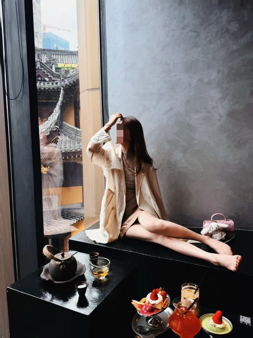 A young woman with her face blurred sitting on a black counter in what appears to be a tea house, with traditional architecture visible through a window and various desserts arranged in the foreground.