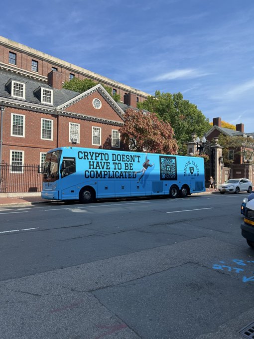 A light blue bus parked on a city street displaying the advertisement 'CRYPTO DOESN'T HAVE TO BE COMPLICATED' with a QR code for 'Phantom Wallet,' with a historic brick building in the background.