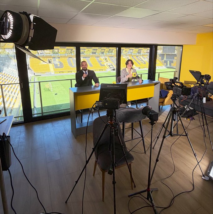 A behind-the-scenes view of a broadcast studio overlooking a stadium with yellow seats, where two presenters sit at a desk with multiple cameras and lighting equipment set up around them.