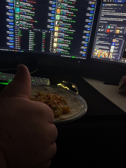 A hand gives a thumbs-up in front of a computer monitor displaying what appears to be a stock or crypto trading interface with green and red numbers, alongside a plate of food and a gaming mouse.