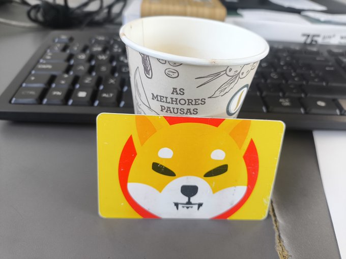 A Shiba Inu-themed Tangem crypto card rests on a desk beside a paper coffee cup that reads "AS MELHORES PAUSAS," with a keyboard in the background—matching the caption's celebration of the card paired with a morning coffee funded via Tangem Pay.